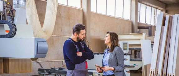 Mann und Frau unterhalten sich in einer Werkstatt mit Maschinen und Holzmaterialien im Hintergrund.