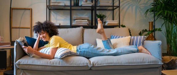 Person mit lockigem Haar liegt auf grauem Sofa, liest Buch, trägt gelbes Shirt und Jeans, modernes Wohnzimmer mit Pflanzen und Regalen.