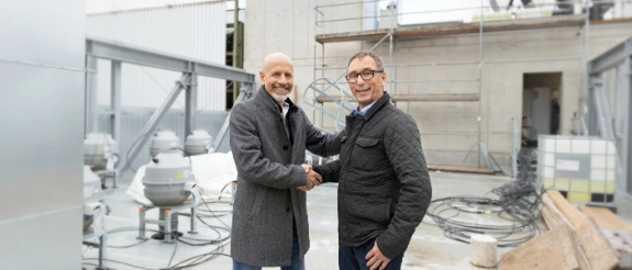 Zwei Männer schütteln sich die Hand auf einer Baustelle vor Industrieanlagen und Gerüsten.
