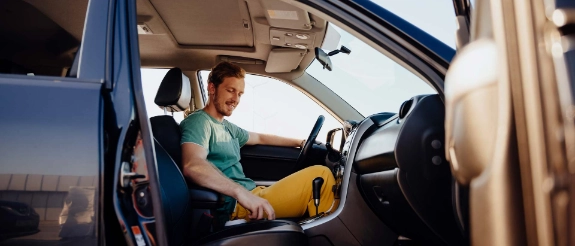 Mann sitzt am Steuer eines Autos mit offener Fahrertür, hält Lenkrad, sonniges Wetter, helles Interieur