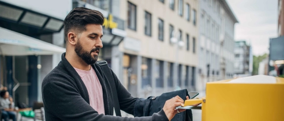 Mann mit Bart wirft Müll in gelben Behälter auf urbaner Straße mit Gebäuden im Hintergrund
