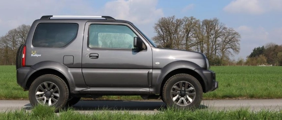 Silberner Suzuki Jimny SUV Geländewagen in Seitenansicht auf grüner Wiese mit Bäumen im Hintergrund