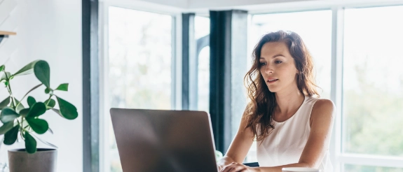 Frau arbeitet konzentriert am Laptop an hellem Holztisch im modernen Büro mit Pflanze und großem Fenster