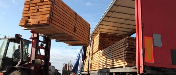Gabelstapler lädt Holzbalken auf Lastkraftwagen, Baustoffhandel mit gestapelten Holzbrettern im Hintergrund