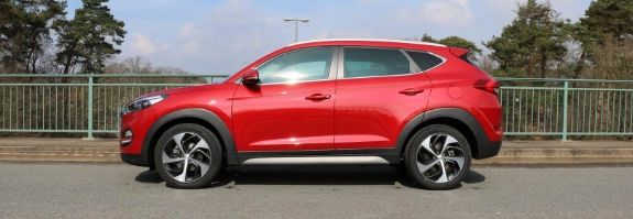 Roter SUV steht seitlich auf Parkplatz mit Metallzaun und Bäumen im Hintergrund