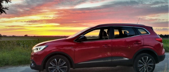 Roter SUV parkt bei Sonnenuntergang auf Landstraße mit dramatischem Himmel und Grünland