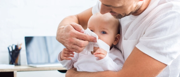 Vater hält Baby im Arm und putzt ihm mit Tuch aus der Hausapotheke für Babys die Nase