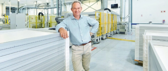 Mann in blauem Hemd lehnt an Metallplatte in Industriehalle mit gelben Schutzvorrichtungen und Maschinen im Hintergrund