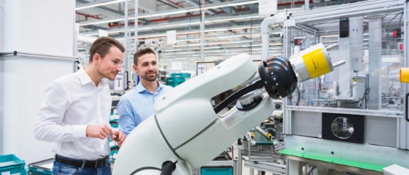 Zwei Männer inspizieren einen weißen Industrieroboter in einer modernen Fabrikhalle mit Werkbänken und Maschinen.
