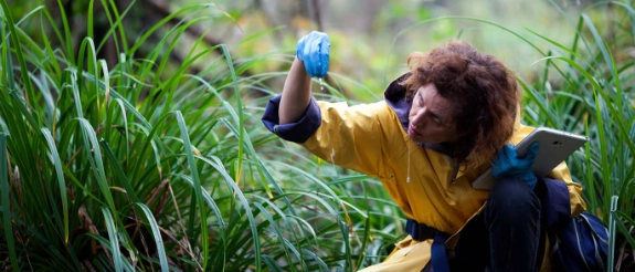 Person mit lockigem Haar in gelbem Oberteil hebt Hand mit blauem Objekt in grüner Natur in die Luft.