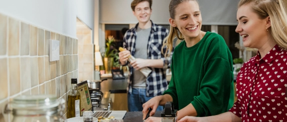 Drei Personen kochen gemeinsam in einer modernen Küche, eine Frau in grünem Pulli bereitet Essen zu.