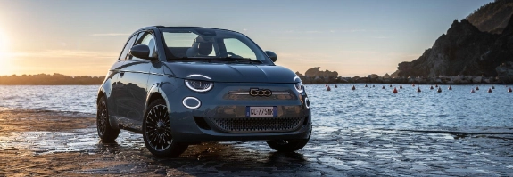 Blauer Fiat 500e am Strand bei Sonnenuntergang, im Hintergrund Wasser und Segelboote