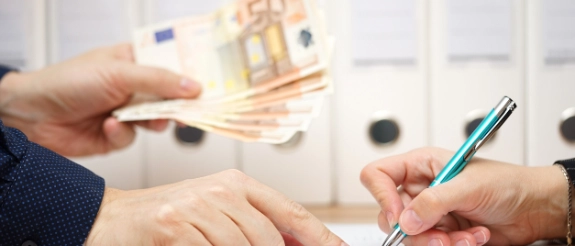 Zwei Personen unterzeichnen einen Vertrag, während eine Hand mehrere Euro-Scheine hält, im Hintergrund Ordner.