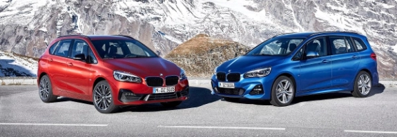 Zwei BMW Fahrzeuge in Rot und Blau stehen sich auf einer Bergstraße gegenüber, schneebedeckte Alpen im Hintergrund