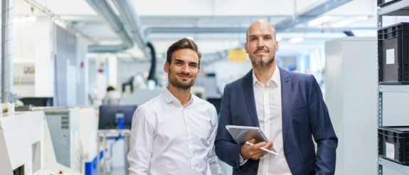 Zwei Geschäftsmänner in Business-Kleidung stehen in moderner Fabrik- oder Büroumgebung, einer hält Tablet