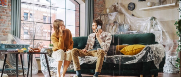 Zwei Frauen renovieren Wohnzimmer, halten Farbroller, bunte Farbeimer auf Boden, helles modernes Apartment mit großem Fenster.