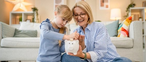 Großmutter und Enkelin werfen Münz-Geld in ein Sparschwein als Symbol für das Sparen für Kinder.