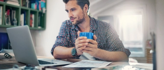 Mann im karierten Hemd hält blaue Tasse und schaut auf Dokumente am Schreibtisch
