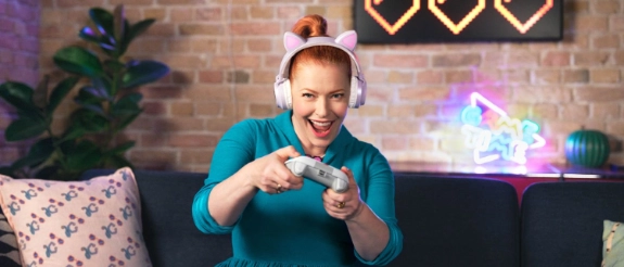 Lachende Frau in blauem Kleid sitzt auf Sofa, macht Gaming-Gesten, Ziegelwand mit Neon-Herzen und Pflanze im Hintergrund