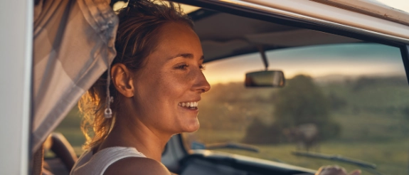 Lächelnde Frau schaut aus Autofenster auf grüne Landschaft bei Sonnenuntergang