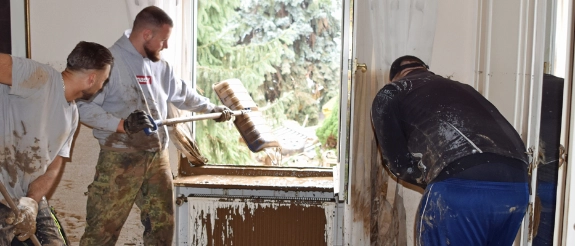 Person reinigt Fensterrahmen mit Bürste, Wald im Hintergrund, alte weiße Fensterbank mit Farbabplatzungen sichtbar.