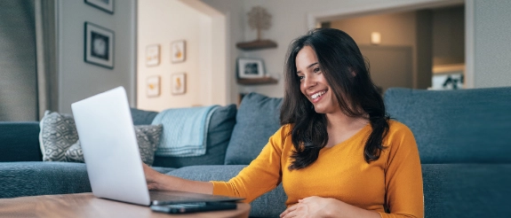 Frau in gelbem Shirt arbeitet lächelnd am Laptop auf dem Couchtisch, gemütliches Wohnzimmer mit grauem Sofa im Hintergrund.