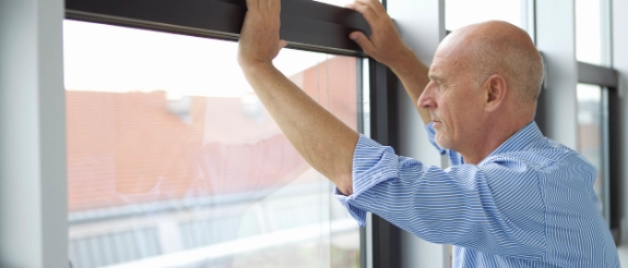 Mann in blauem Hemd bringt schwarze Fensterfolie an modernem Fenster an, Stadtblick im Hintergrund