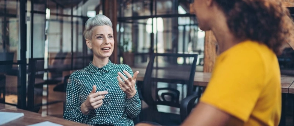 Zwei Frauen unterhalten sich an einem Tisch in einem modernen Büro mit großen Fenstern im Hintergrund.