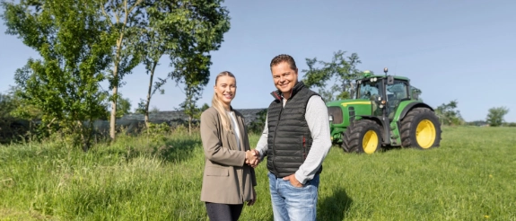 Zwei Personen schütteln Hände auf einer Wiese, im Hintergrund ein grüner Traktor und Bäume