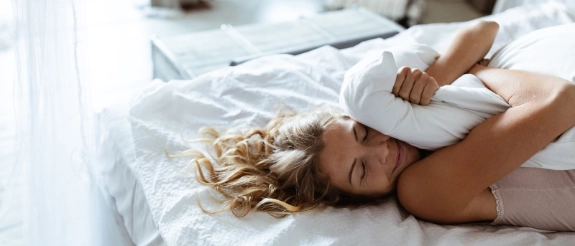 Frau mit lockigem Haar liegt auf weißem Bett und liest Zeitung in hellem Schlafzimmer