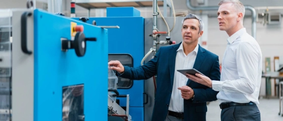 Zwei Geschäftsmänner inspizieren eine blaue Industriemaschine in einer Fabrik, einer hält ein Tablet