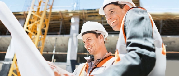 Zwei Bauarbeiter mit Schutzhelmen und Warnwesten besprechen Baupläne auf einer Baustelle.