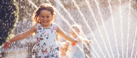 Lachendes Mädchen in buntem Kleid springt durch Wasserstrahlen eines Gartensprengers an sonnigem Sommertag