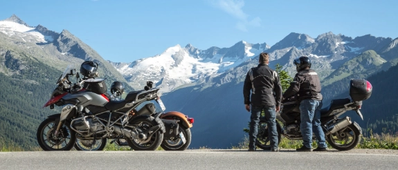 2 Freunde sind mit dem Motorrad im Ausland unterwegs. Sie haben angehalten und blicken auf die Berge