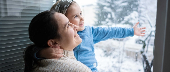 Eine Mutter hat ihr kleines Kind auf dem Arm und beide schauen aus dem Fenster. 