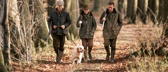 Drei Jäger mit Gewehren und Hund gehen durch herbstlichen Wald mit Laubstreu