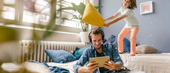 Vater hört Music auf dem Tablet und Kind tobt auf dem Sofa und spielt Kissenschlacht.