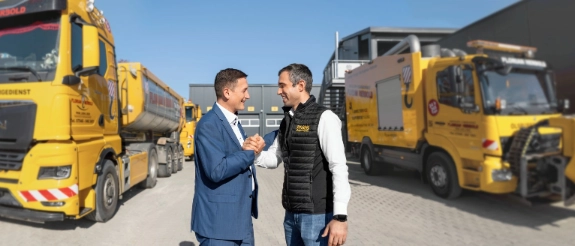 Zwei Männer schütteln sich die Hand vor gelben Lastkraftwagen auf einem Logistik-Betriebshof.