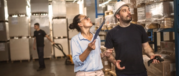 Zwei Personen inspizieren Lagerhalle mit Clipboard und Bauhelm, Lagerbestände im Hintergrund sichtbar.