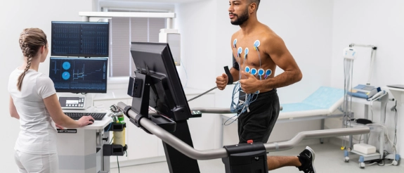 Mann auf Laufband bei Belastungs-EKG, Frau überwacht Vitaldaten am Monitor in medizinischer Praxis.