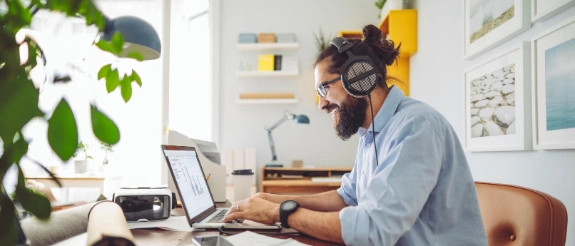 Mann mit Brille und Kopfhörern arbeitet am Laptop in modernem Home Office mit Pflanzen und gelben Regalen