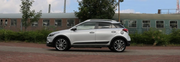 Weißer SUV parkt auf Ziegelsteinen vor Wohngebäuden mit Bäumen und bewölktem Himmel