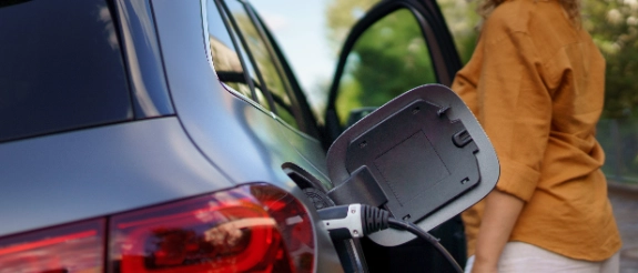 Elektroauto-Ladekabel an Fahrzeug angeschlossen, Person im orangenen Shirt daneben, grüne Umgebung sichtbar