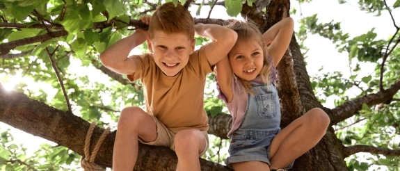 Zwei lachende Kinder sitzen auf einem Baumast im Freien, umgeben von grünem Laub und Sonnenlicht