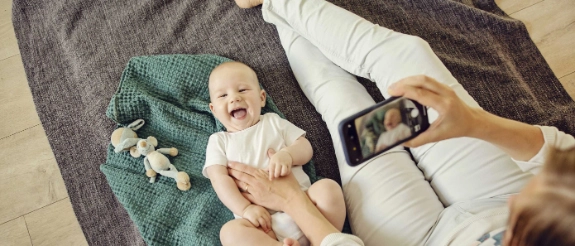 Mutter fotografiert lachendes Baby auf grauer Decke mit Smartphone, Nahaufnahme von oben