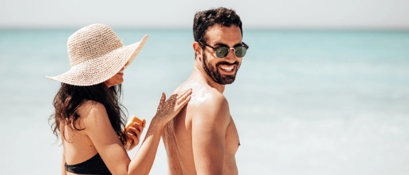 Paar am Strand trägt Sonnenschutz auf: Frau mit Strohhut, Mann mit Sonnenbrille, türkisblaues Meer im Hintergrund
