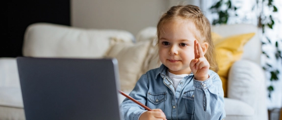 Kleines Mädchen sitzt am Schreibtisch vor Laptop, hält Buntstift, lächelt in die Kamera, Notizbuch und Farbstifte sichtbar.