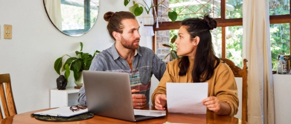 Mann und Frau sitzen am Tisch mit Laptop und Papieren, diskutieren gemeinsam in hellem Büro mit Pflanzen.