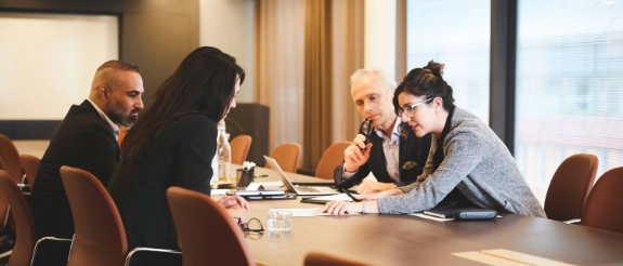 Geschäftstreffen mit vier Personen an Konferenztisch, Mann spricht ins Mikrofon, modernes Büro mit Panoramafenstern.