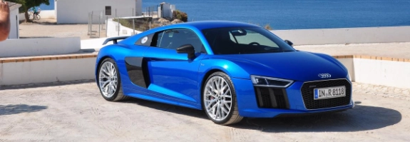 Blauer Audi R8 Sportwagen an Küstenstrand mit weißem Haus und Mittelmeer im Hintergrund